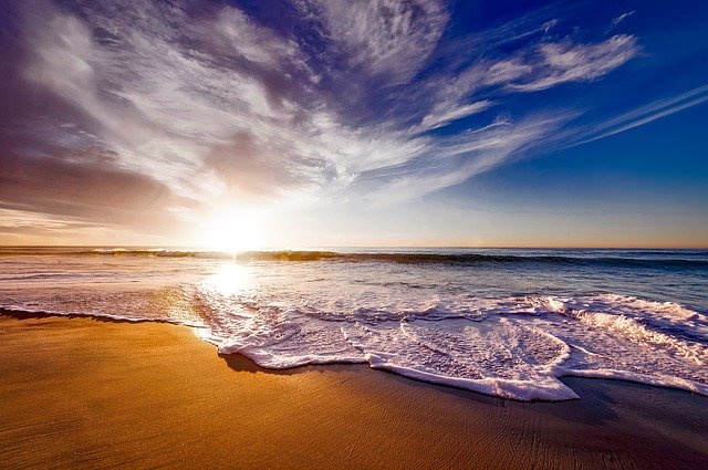 Photograph of the sunset over the Pacific Ocean. This is a placid beach with golden sand and gentle waves. The sun is touching the horizon and as it illuminates the wispy clouds from underneath and reflects off the ripples in the water. There is a faint line of orange on the horizon with a deep blue sky above.